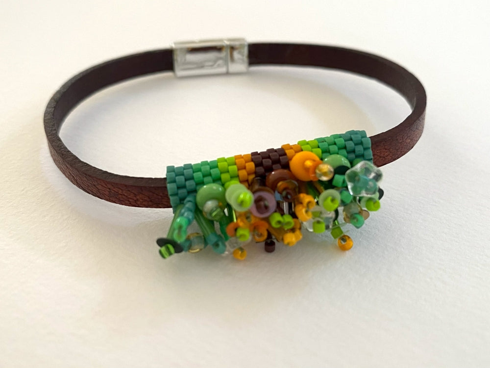 A dark brown leather bracelet featuring a colorful beaded floral centerpiece in shades of green, yellow, orange, and brown, secured with a silver clasp on a white background.