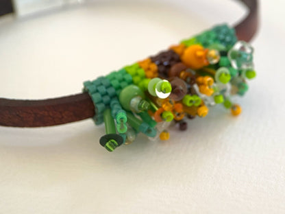 A close-up of a handmade beaded and woven bracelet featuring a brown leather band, green seed beads, and colorful accents of orange, yellow, and white.