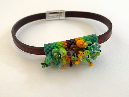 A dark brown leather bracelet featuring a colorful beaded floral centerpiece in shades of green, yellow, orange, and brown, secured with a silver clasp on a white background.